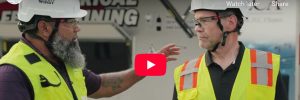 Two men in high visibility vests, hardhats, and safety glasses talking in front of the NECA safety training trailer.